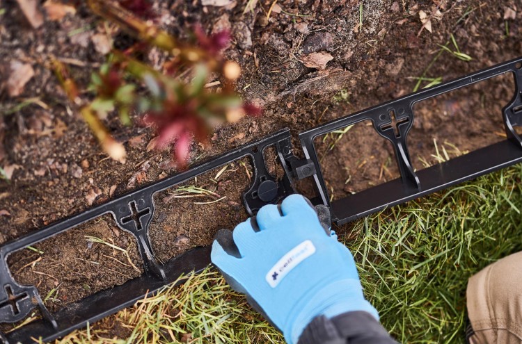 Ręka w niebieskiej rękawicy montująca czarne proste obrzeże ogrodowe UNIBORD na trawniku i ziemi